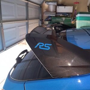 Könnte beinhalten: Ein Heckspoiler aus Kohlefaser mit den blauen Buchstaben "RS" auf einem blauen Auto. Der Spoiler hat ein strukturiertes Webmuster und ist am Heck des Fahrzeugs montiert. Das Auto steht in einer Garage.