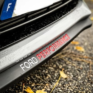 Op de afbeelding: Close-up van de voorbumper van een auto met de tekst "FORD PERFORMANCE" in rode en witte letters. De bumper is zwart en grijs, met een strak, aerodynamisch ontwerp. Een Frans kenteken is zichtbaar op de achtergrond.