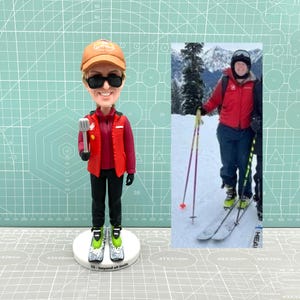May include: A custom bobblehead figurine of a person wearing a red jacket, black pants, and a brown hat with a logo. The figurine is holding a spatula in their right hand. The background is a green grid pattern. The figurine is standing on a white base with the text "55 - beyond all limits".
