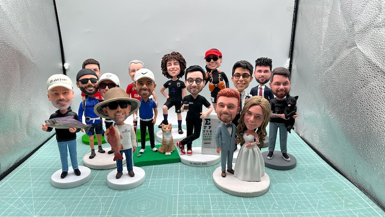 May include: A collection of custom bobblehead figurines, each depicting a person in various outfits and poses. Some hold objects like fish, a cat, and a coffee cup. The figurines are on white circular bases.