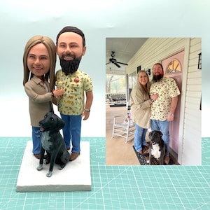 May include: A custom-made figurine set of a couple and their dog. The figurines are made of clay and are painted in realistic colours. The couple are wearing casual clothing and the dog is sitting next to them. The figurines are standing on a white base.