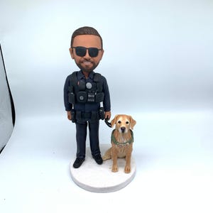 May include: A figurine of a police officer with sunglasses and a golden retriever dog. The officer wears a dark blue uniform and tactical vest. The dog wears a green vest with "POLICE" on it. The figurine is on a white circular base.