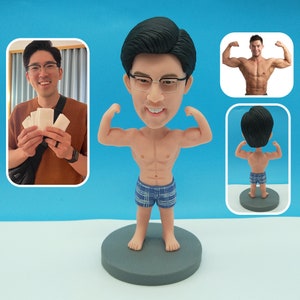 以下が含まれることがあります： 筋肉質の体格の人物のカスタムフィギュアで、青と白の格子縞のショートパンツを着用しています。フィギュアは腕を曲げてポーズをとっており、たくましい体格を強調しています。フィギュアは灰色の台座の上に立っています。