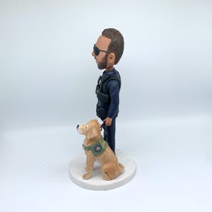 May include: A figurine of a man in a dark blue uniform and sunglasses, standing next to a golden retriever wearing a green vest. The dog is on a leash. The figurine is on a white circular base.