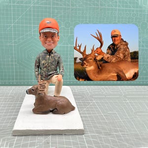 May include: A figurine of a hunter kneeling next to a deer. The hunter wears a camouflage jacket and orange hat. The deer is brown with antlers. A smaller image shows a hunter with a deer.