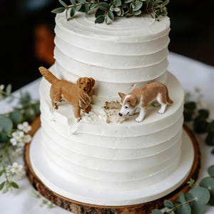 Könnte beinhalten: Eine zweistöckige weiße Hochzeitstorte mit strukturierter Glasur, verziert mit zwei kleinen Hundefiguren. Der Kuchen steht auf einem Holzsockel und ist mit Grünzeug verziert.