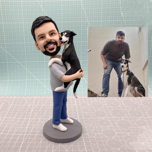 May include: A custom bobblehead figurine of a person holding a black and white dog. The figurine is on a gray base. The person is wearing a gray shirt, blue pants, and white shoes. The dog is licking the person's face.