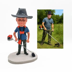 May include: A figurine of a person wearing a black hat, blue shirt, and orange harness, holding a string trimmer. The figurine is on a gray base, with a smaller image of a person using a string trimmer in a grassy field.