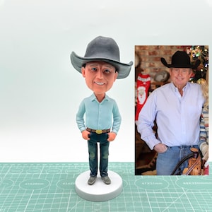 May include: A custom bobblehead figurine of a person wearing a blue button-down shirt, blue jeans, and a gray cowboy hat. The figurine is standing on a white base. The person is smiling and has a friendly expression.