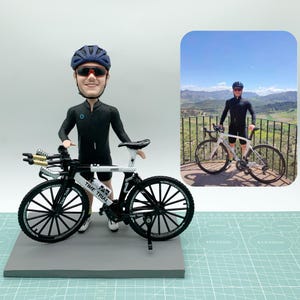 Peut inclure: Une figurine bobblehead personnalisée représentant un cycliste portant un casque bleu foncé, des lunettes de soleil teintées de rouge et un maillot de cyclisme noir avec un short, à côté d'un vélo de contre-la-montre miniature blanc et noir.