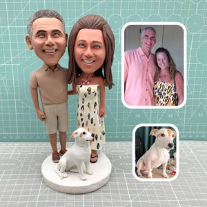 May include: A custom bobblehead figurine featuring a couple and a dog. The couple is dressed in casual attire, and the dog is white with brown markings. The figurine is on a round base, with a photo of the couple and the dog.