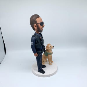 May include: A figurine of a police officer with a golden retriever dog. The officer wears a dark blue uniform, sunglasses, and a tactical vest. The dog has a green harness. The figurine is on a white circular base.