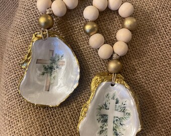 Puede incluir: Dos adornos decorativos de concha de ostra. Cada concha está pintada de blanco con un borde dorado y presenta un diseño de cruz con follaje verde. Los adornos están conectados a una cadena de cuentas de madera, con cuentas doradas.