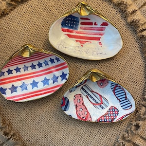 May include: Three decorative seashell dishes with patriotic designs. One features the American flag and the text "Home Sweet Home." Another has red and white stripes with blue stars. The third has a pattern of red, white, and blue flip-flops.