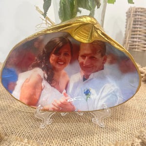 Könnte beinhalten: Eine goldbemalte Muschel mit einem Foto eines Paares darin. Das Paar lächelt und hält sich an den Händen. Die Muschel steht auf einem durchsichtigen Plastikständer.