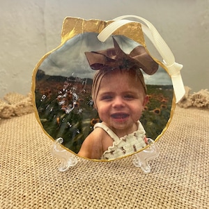May include: A gold-rimmed seashell ornament featuring a photograph of a young child wearing a brown bow and a white dress. The ornament is displayed on a white stand.