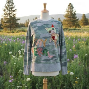 May include: Camo-print sweatshirt with a gray bottom and sleeves. The sweatshirt features a colorful, stitched design of a cat and the words "Say Real". The sweatshirt is displayed on a mannequin in a field of wildflowers.