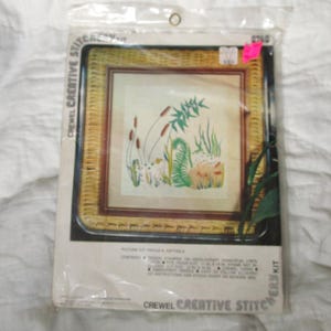 May include: A Crewel Creative Stitchery Kit in a clear plastic package. The kit features a framed needlepoint design of cattails and other marsh plants. The frame is a woven, light brown material. The text "Picture Kit Frogs & Cattails" is visible.