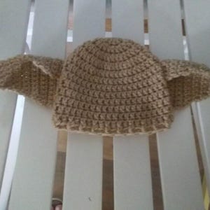 Puede incluir: Un gorro de ganchillo marr&oacute;n claro, con dos grandes extensiones en forma de alas a cada lado. El gorro est&aacute; hecho con un punto texturizado, y las alas tambi&eacute;n est&aacute;n tejidas a ganchillo. El gorro est&aacute; sobre una superficie blanca.