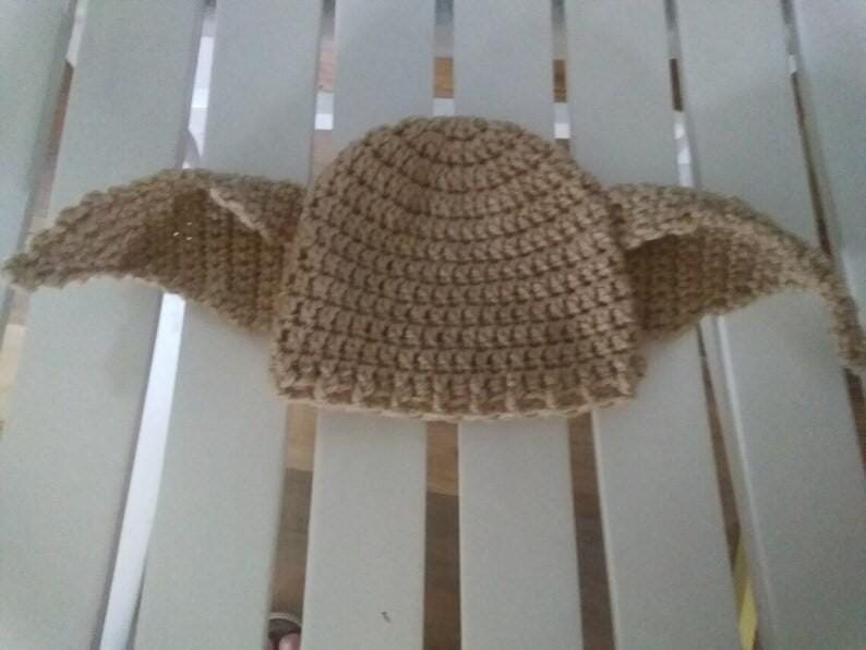 Puede incluir: Un gorro de ganchillo marr&oacute;n claro, con dos grandes extensiones en forma de alas a cada lado. El gorro est&aacute; hecho con un punto texturizado, y las alas tambi&eacute;n est&aacute;n tejidas a ganchillo. El gorro est&aacute; sobre una superficie blanca.