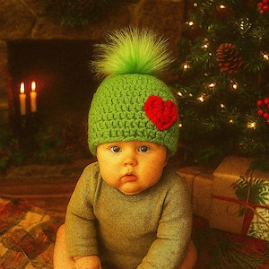 May include: A baby wearing a green knitted hat with a red heart applique and a green fluffy pom-pom. The baby is also wearing a green long-sleeved shirt. The background includes a Christmas tree, candles, and a fireplace.