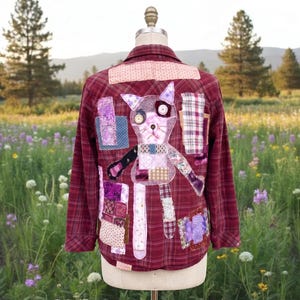 May include: A burgundy plaid shirt with a patchwork cat design on the back. The cat is made of various fabric scraps in shades of purple, pink, and beige. The shirt is displayed on a mannequin in a field of wildflowers.