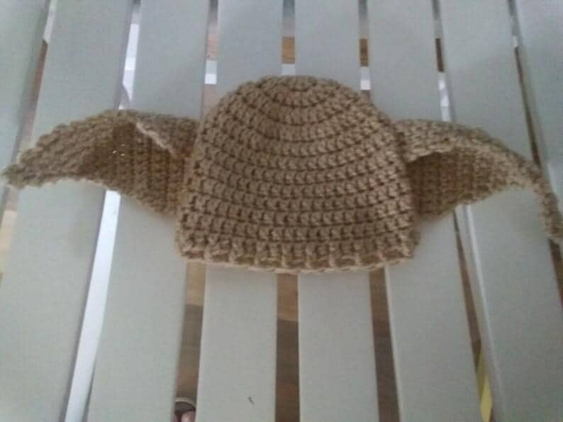 Puede incluir: Un gorro de ganchillo de color marr&oacute;n claro, con dos grandes extensiones en forma de alas a cada lado. El gorro est&aacute; hecho con un punto texturizado, y las alas tambi&eacute;n est&aacute;n hechas a ganchillo. El gorro est&aacute; sobre una superficie blanca.