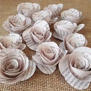 May include: A collection of paper roses crafted from book pages. The roses are a soft, neutral color, with visible text from the book adding texture and detail. The petals are carefully shaped to resemble real roses, creating a unique and artistic floral arrangement.