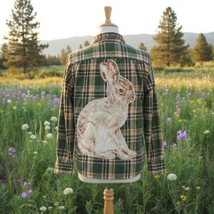May include: A green and brown plaid flannel shirt with a large, detailed illustration of a rabbit on the back. The shirt is displayed on a mannequin in a field of wildflowers.