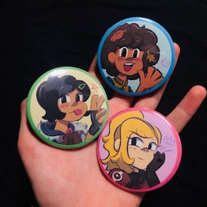 Puede incluir: Tres pines redondos con personajes de dibujos animados. El primer pin tiene un fondo verde con un personaje femenino con cabello castaño y una camisa verde. El segundo pin tiene un fondo azul con un personaje femenino con cabello castaño y una camisa marrón. El tercer pin tiene un fondo rosa con un personaje femenino con cabello rubio y una camisa roja.