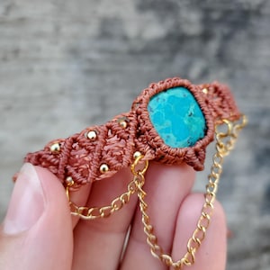 May include: A macrame bracelet featuring a central rectangular turquoise stone set in brown cord. The band is accented with small gold-toned beads and features two delicate gold-toned metal chains hanging from the sides of the stone setting.