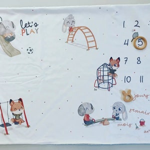 Può includere: Una copertina bianca per i traguardi del bambino con un tema di parco divertente. Presenta illustrazioni di animali su altalene, scivoli e un'altalena. La copertina ha una tabella di numeri da 1 a 12, con spazi per i marcatori di legno per tracciare la crescita di un bambino in giorni, settimane, mesi e anni. Il testo "Let's Play" è stampato sulla copertina.