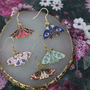 May include: Four pairs of gold-tone metal moth earrings with colorful enamel wings. The earrings feature pink, blue, and light green wings with intricate details. The earrings are displayed on a decorative, gold-edged, oval-shaped tray.