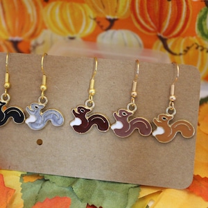 May include: Five pairs of dangle earrings featuring squirrel designs in black, gray, brown, and tan with white bellies and gold-colored hooks. The earrings are displayed on a brown card, with a fall-themed background.