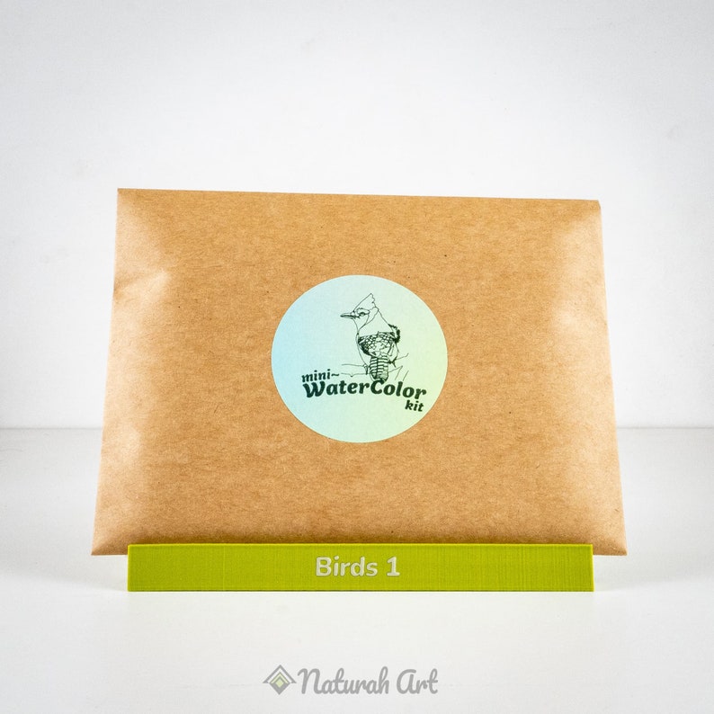 Puede incluir: Un sobre de papel marr&oacute;n con una pegatina que dice "mini-Watercolor kit" y un dibujo de un arrendajo azul. El sobre est&aacute; sobre una barra verde que dice "Birds 1" y un logotipo que dice "Naturah Art".