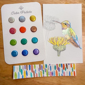 Pode incluir: Uma paleta de cores em aquarela com 12 pontos de tinta metálica em várias cores, uma pintura em aquarela de um beija-flor em uma flor amarela e um marcador de aquarela com detalhes metálicos coloridos. A paleta é rotulada como "Color Palette".