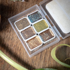 May include: A clear plastic case containing six metallic watercolor paints in various shades of gold, bronze, and teal. The paints are in small, rectangular pans with white borders. The case is open, revealing the paints against a wooden surface.