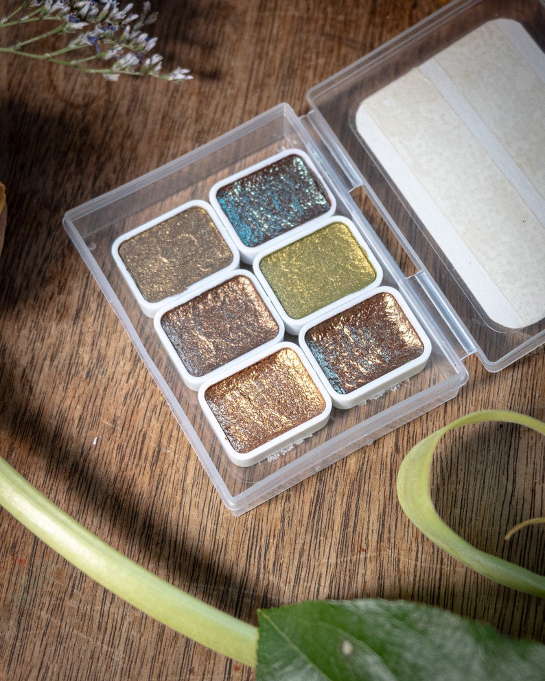 May include: A clear plastic case containing six metallic watercolor paints in various shades of gold, bronze, and teal. The paints are in small, rectangular pans with white borders. The case is open, revealing the paints against a wooden surface.