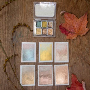 May include: A clear plastic watercolor palette with six metallic shades, displayed with six watercolor swatches on white paper. The palette is open, revealing shades of gold, bronze, and green. A dried leaf and sprig of berries are also present.