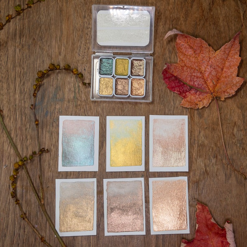 May include: A clear plastic watercolor palette with six metallic shades, displayed with six watercolor swatches on white paper. The palette is open, revealing shades of gold, bronze, and green. A dried leaf and sprig of berries are also present.