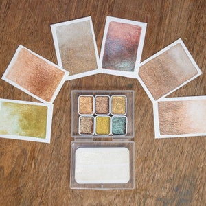 May include: A watercolor paint set with six metallic shades in a clear plastic case, surrounded by watercolor swatches. The swatches display various metallic hues, including gold, bronze, and green, on white paper. The background is a wooden surface.