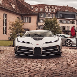 Könnte beinhalten: Ein silberner Bugatti Chiron Pur Sport, ein Hochleistungs-Sportwagen, ist vor einem Gebäude mit einem Kopfsteinpflaster-Fahrweg geparkt. Das Auto hat einen schwarzen Kühlergrill und einen schwarzen Streifen, der über die Mitte der Motorhaube verläuft.