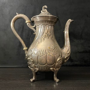 May include: A silver teapot with an ornate design and a lid. The teapot has a curved spout and a handle.
