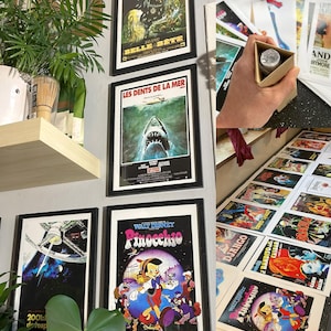May include: A collection of framed film posters, including "Belle et Bete", "Les Dents de la Mer", and "Pinocchio", adorn a wall. More posters are spread out, and a person holds a small box. The posters are in black frames.