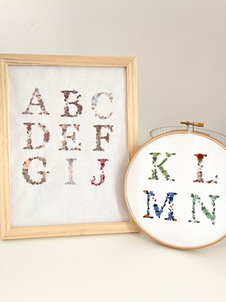 Pu&ograve; includere: Due cerchi da ricamo con le lettere da A a N ricamate in motivi floreali. Le lettere da A a J sono in una cornice di legno, e le lettere da K a N sono in un cerchio separato. Le lettere sono ricamate con fiori di diversi colori.