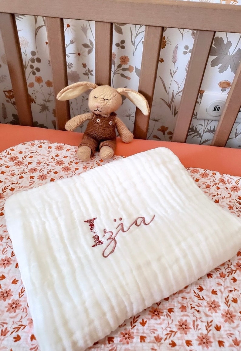 Pu&ograve; includere: Coperta per neonato in mussola bianca con il nome "Izja" ricamato in filo rosa. La coperta &egrave; piegata e appoggiata su un lenzuolo da culla floreale rosa e bianco.
