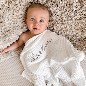 Pu&ograve; includere: Una copertina per neonati in mussola bianca con il nome "Marlon" ricamato in filo marrone. La copertina &egrave; avvolta intorno a un bambino sdraiato su una superficie pelosa beige.