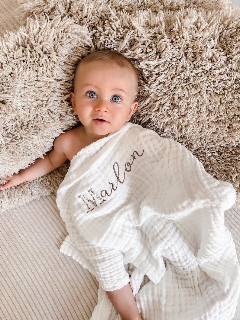 Pu&ograve; includere: Una copertina per neonati in mussola bianca con il nome "Marlon" ricamato in filo marrone. La copertina &egrave; avvolta intorno a un bambino sdraiato su una superficie pelosa beige.