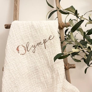 Pu&ograve; includere: Coperta per neonati in cotone bianco con una ghirlanda di fiori e il nome "Olympe" ricamato in filo marrone.