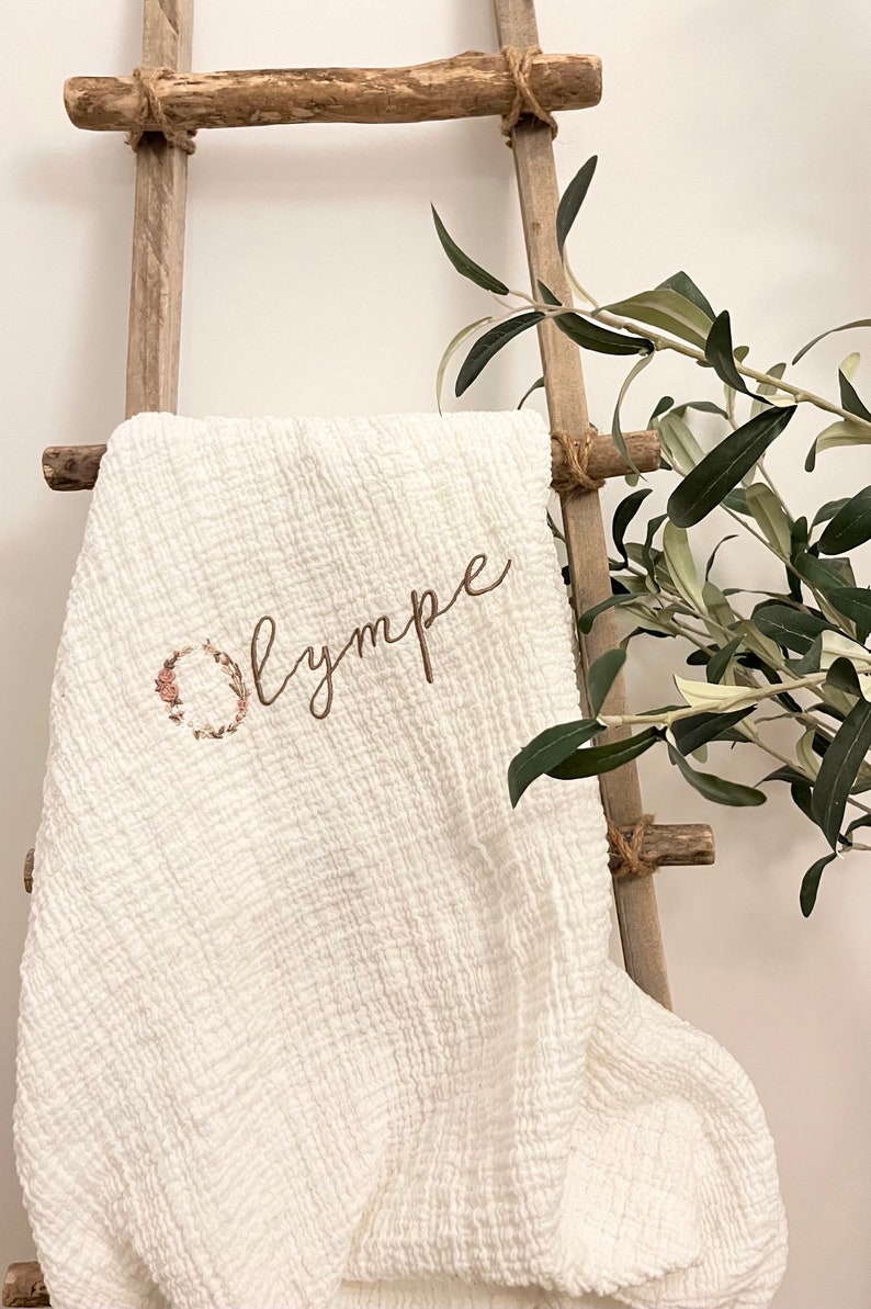 Pu&ograve; includere: Coperta per neonati in cotone bianco con una ghirlanda di fiori e il nome "Olympe" ricamato in filo marrone.
